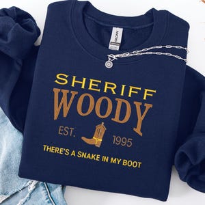 Puede incluir: Sudadera azul marino con el texto "SHERIFF WOODY" en letras doradas y marrones. El dise&ntilde;o incluye una bota de vaquero y la frase "THERE'S A SNAKE IN MY BOOT". Un collar plateado con un colgante de sol est&aacute; sobre la sudadera.