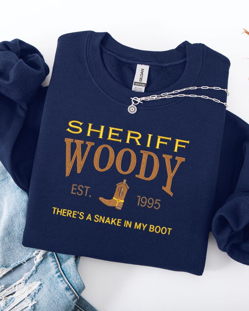 Puede incluir: Sudadera azul marino con el texto "SHERIFF WOODY" en letras doradas y marrones. El dise&ntilde;o incluye una bota de vaquero y la frase "THERE'S A SNAKE IN MY BOOT". Un collar plateado con un colgante de sol est&aacute; sobre la sudadera.