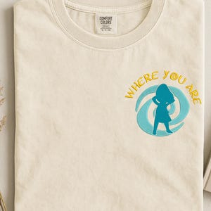 Może przedstawiać: Kremowa koszulka z haftowanym napisem "WHERE YOU ARE" w kolorze ż&oacute;łtym, otaczającym niebieską sylwetkę osoby. Koszulka ma okrągły dekolt i metkę z napisem "COMFORT COLORS".