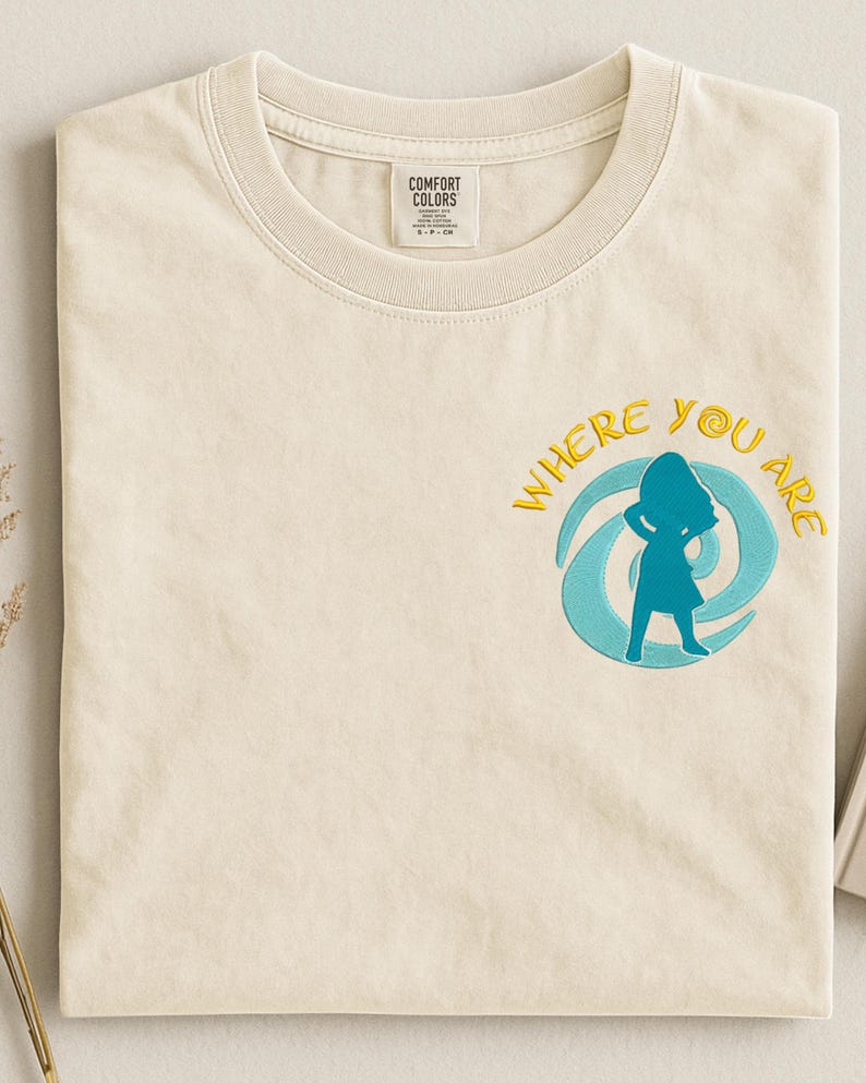 Może przedstawiać: Kremowa koszulka z haftowanym napisem "WHERE YOU ARE" w kolorze ż&oacute;łtym, otaczającym niebieską sylwetkę osoby. Koszulka ma okrągły dekolt i metkę z napisem "COMFORT COLORS".