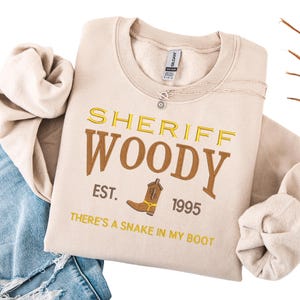Puede incluir: Sudadera beige con bordado amarillo y marr&oacute;n "SHERIFF WOODY" y un dibujo de bota de vaquero. El texto "EST. 1995" y "THERE'S A SNAKE IN MY BOOT" tambi&eacute;n est&aacute;n bordados. Un collar plateado est&aacute; sobre la sudadera.