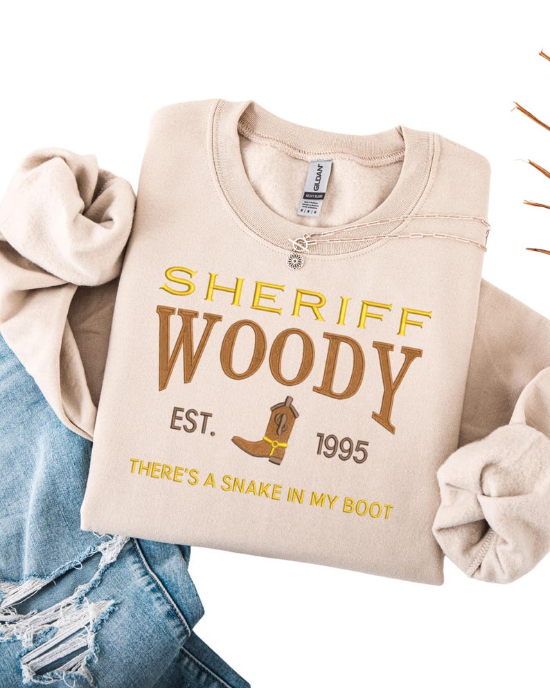 Puede incluir: Sudadera beige con bordado amarillo y marr&oacute;n "SHERIFF WOODY" y un dibujo de bota de vaquero. El texto "EST. 1995" y "THERE'S A SNAKE IN MY BOOT" tambi&eacute;n est&aacute;n bordados. Un collar plateado est&aacute; sobre la sudadera.