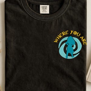 Może przedstawiać: Czarny t-shirt z okrągłym dekoltem i haftowanym wzorem. Wz&oacute;r przedstawia niebieską sylwetkę w niebiesko-ż&oacute;łtym wirze z napisem "WHERE YOU ARE". Koszulka ma metkę "COMFORT COLORS".