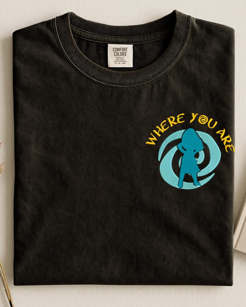 Może przedstawiać: Czarny t-shirt z okrągłym dekoltem i haftowanym wzorem. Wz&oacute;r przedstawia niebieską sylwetkę w niebiesko-ż&oacute;łtym wirze z napisem "WHERE YOU ARE". Koszulka ma metkę "COMFORT COLORS".