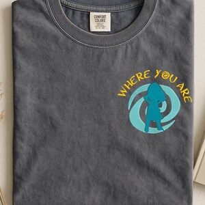 Może przedstawiać: Szara koszulka z okrągłym dekoltem. Koszulka ma napis "WHERE YOU ARE" w kolorze ż&oacute;łtym, z turkusową grafiką przedstawiającą osobę wewnątrz wiru. Mała metka jest widoczna przy dekolcie.