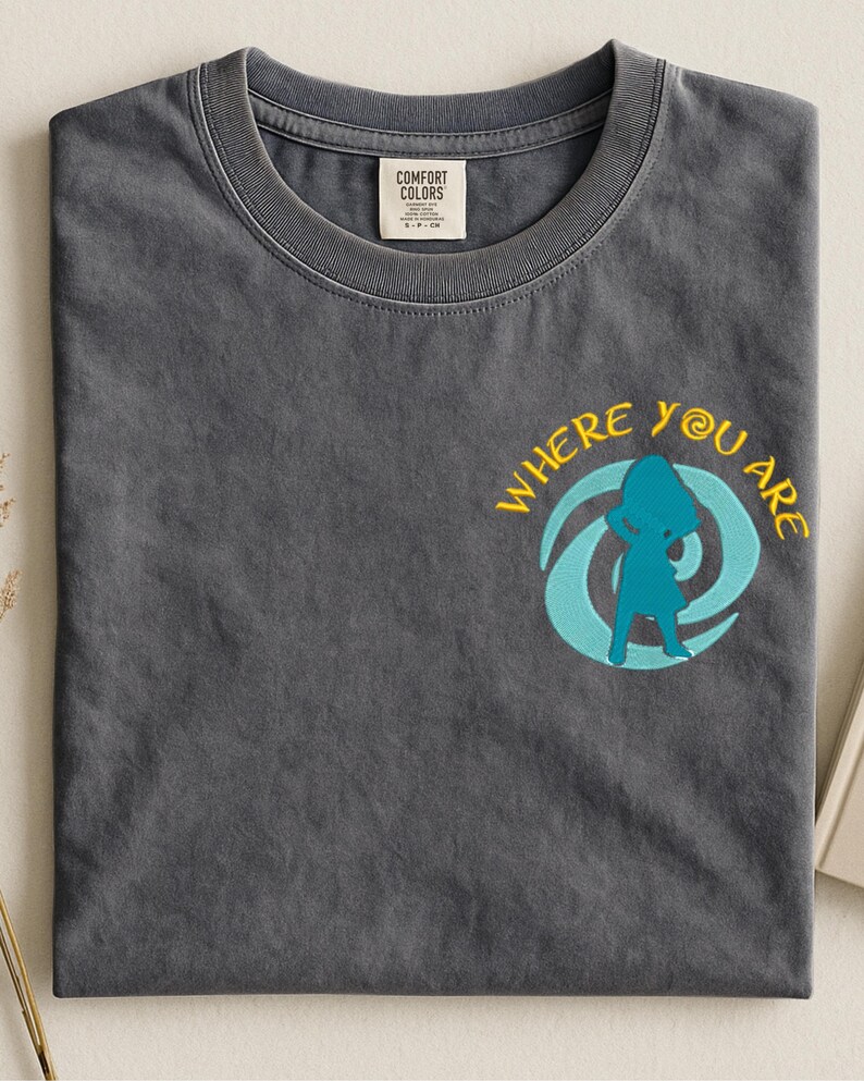 Może przedstawiać: Szara koszulka z okrągłym dekoltem. Koszulka ma napis "WHERE YOU ARE" w kolorze ż&oacute;łtym, z turkusową grafiką przedstawiającą osobę wewnątrz wiru. Mała metka jest widoczna przy dekolcie.