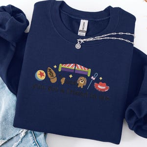 May include: Navy blue sweatshirt with embroidered Toy Story-themed graphics. Features a ball, Buzz Lightyear's wings, a dog, a cowboy hat, and the phrase "you got a friend in me". A silver necklace with a sun pendant is draped across the sweatshirt.