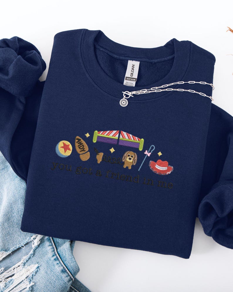 May include: Navy blue sweatshirt with embroidered Toy Story-themed graphics. Features a ball, Buzz Lightyear's wings, a dog, a cowboy hat, and the phrase "you got a friend in me". A silver necklace with a sun pendant is draped across the sweatshirt.