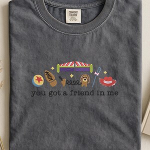 May include: Dark gray Comfort Colors t-shirt with embroidered design. Features a ball, boot, circus tent, dog, lasso, and cowboy hat. The text "you got a friend in me" is embroidered below the design. The shirt has a crew neck.