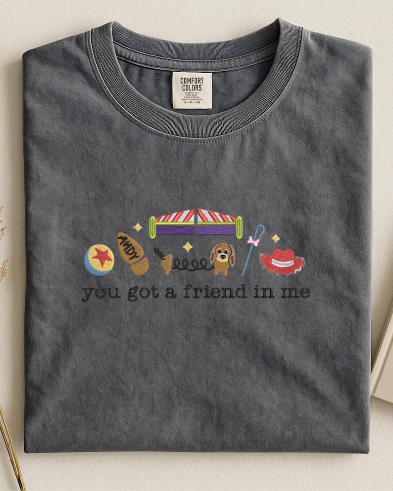 May include: Dark gray Comfort Colors t-shirt with embroidered design. Features a ball, boot, circus tent, dog, lasso, and cowboy hat. The text "you got a friend in me" is embroidered below the design. The shirt has a crew neck.