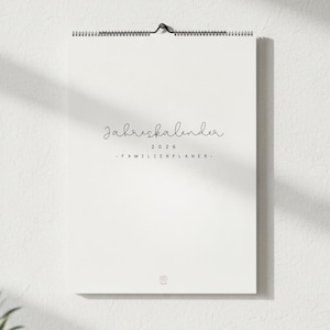 Könnte beinhalten: Ein weißer Wandkalender mit Spiralbindung und schwarzem Metallhaken. Der Kalender zeigt den Text "Jahreskalender 2026 - Familienplaner-" in handschriftlicher Schrift. Unten befindet sich ein kleines Logo.