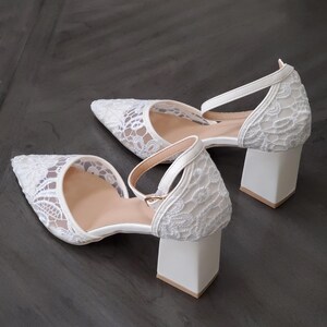 May include: A pair of white lace heels with pointed toes and ankle straps. The heels have a block heel design and a neutral-colored insole. The shoes are set against a dark gray background.