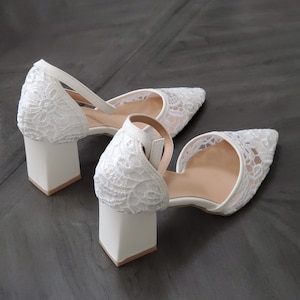 May include: A pair of white lace-covered heels with a pointed toe and a block heel. The shoes have a strap around the ankle and a cutout design on the sides. The heels are approximately 3 inches tall.