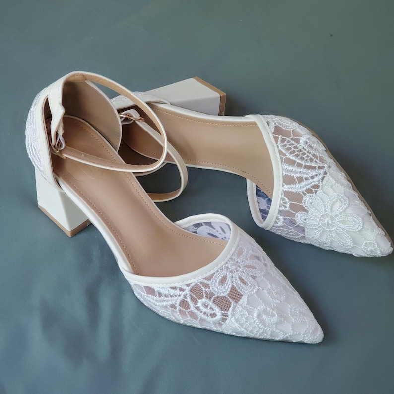 May include: A pair of white lace-covered heels with pointed toes and ankle straps. The shoes have a block heel and a neutral-colored insole. The lace design features floral patterns.