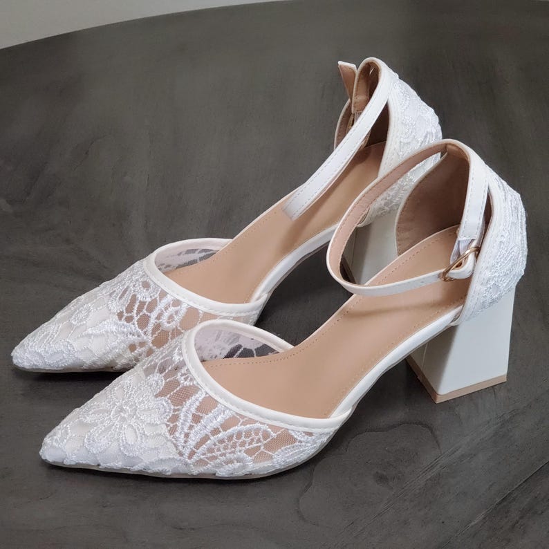 May include: A pair of white lace-covered heels with pointed toes and ankle straps. The heels have a block heel design and a neutral-colored insole. The shoes are set against a neutral background.