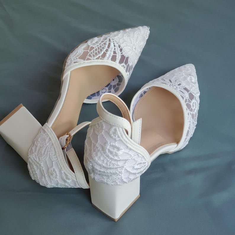May include: A pair of white lace-covered heels with a pointed toe and a block heel. The shoes have an ankle strap with a gold-colored buckle. The heels are approximately 2 inches high.
