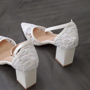 May include: A pair of white lace heels with a pointed toe and a block heel. The heels have a delicate lace overlay and a strap that secures around the ankle. The heel height is approximately 2.5 inches.