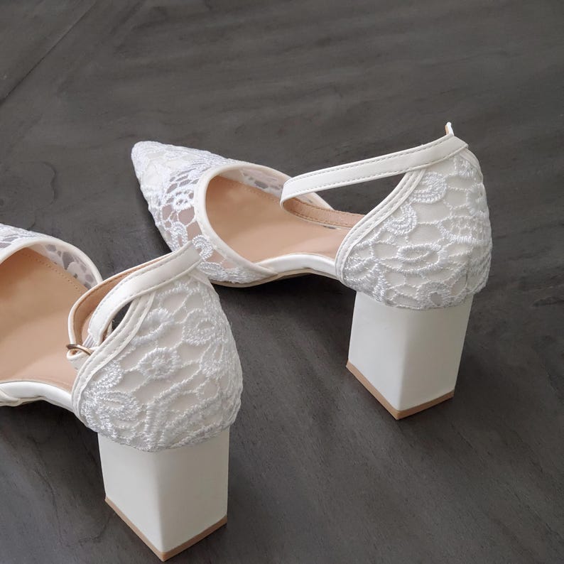 May include: A pair of white lace heels with a pointed toe and a block heel. The heels have a delicate lace overlay and a strap that secures around the ankle. The heel height is approximately 2.5 inches.
