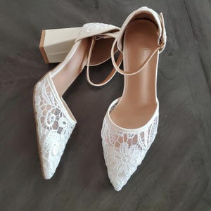 May include: Ivory-colored lace heels with a pointed toe and ankle strap. The heels have a block heel design. The lace detailing features a floral pattern. The shoes are designed for formal occasions.