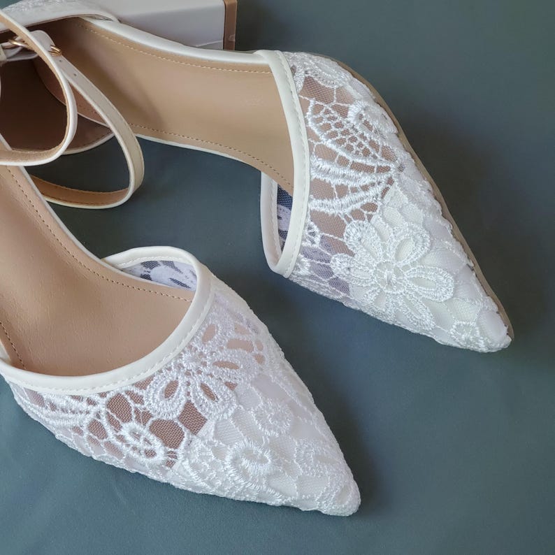May include: White lace pointed-toe heels with ankle straps. The shoes feature a delicate floral lace pattern over a neutral lining. The heels have a low, block heel design.