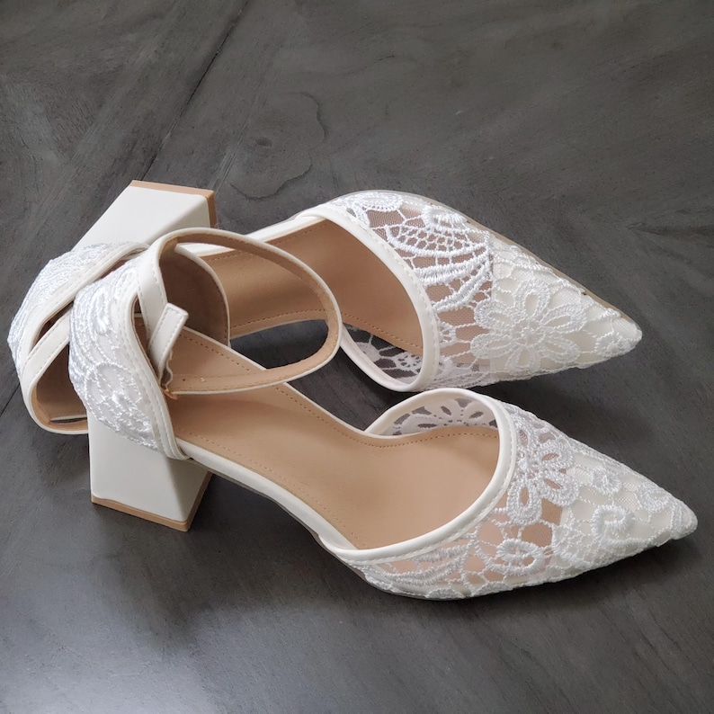 May include: A pair of white lace heels with a pointed toe and ankle strap. The heels have a block heel and a delicate floral lace pattern. The shoes are set against a dark gray background.
