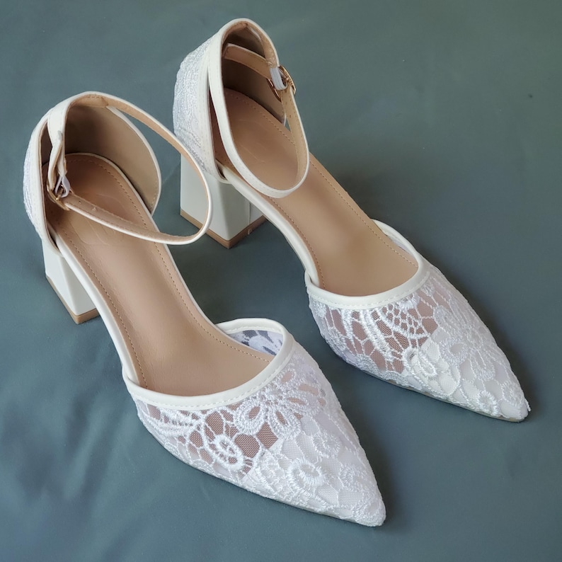 May include: A pair of white lace heels with a pointed toe and ankle strap. The heels have a block heel and a neutral-colored insole. The shoes are set against a solid, muted teal background.