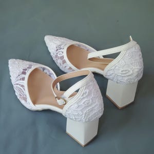 May include: White lace heels with a pointed toe and ankle strap. The heels have a block heel design. The shoes are set against a solid gray-blue background.