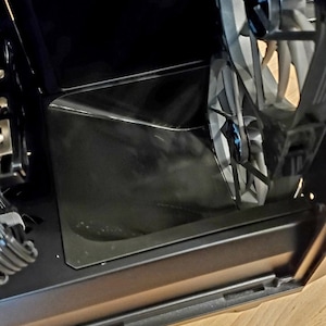 May include: A black computer case with a clear side panel, revealing a silver fan with blue accents. The case is sitting on a light brown wooden surface.