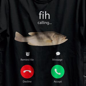 Peut inclure: T-shirt noir avec un motif de poisson et l'inscription "fih calling...". Le design comprend une illustration de poisson et des boutons d'interface téléphonique pour refuser et accepter. Le t-shirt est suspendu à un cintre noir.