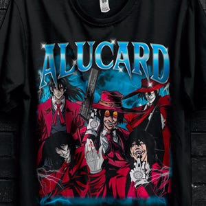Può includere: T-shirt nera con una grafica di personaggi anime in rosso e blu, con la scritta "ALUCARD" in blu. La maglietta presenta anche testo promozionale e una grafica a farfalla. La maglietta è appesa a una gruccia nera.