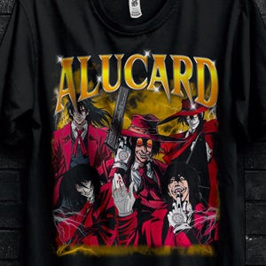 Può includere: T-shirt nera con la scritta "ALUCARD" in oro sopra una grafica di personaggi anime in rosso e nero. La maglietta ha un girocollo ed è appesa a una gruccia nera. L'immagine include anche testo promozionale.
