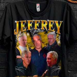 May include: Black t-shirt featuring a graphic of several men with the name "JEFFREY Epstein" in gold lettering. The shirt is hanging on a black hanger against a black brick wall. A butterfly graphic and promotional text are also visible.