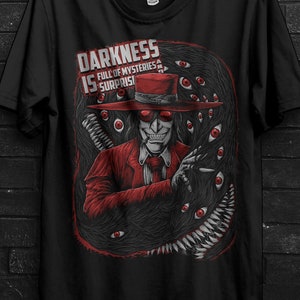May include: Black t-shirt with a graphic of a figure in a red suit and hat, encircled by eyes and a dark, textured backdrop. The text on the shirt says "DARKNESS IS FULL OF MYSTERIES & SURPRISES."