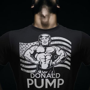 May include: Dark gray t-shirt featuring a muscular illustration of a man with the face of Donald Trump, set against a distressed American flag. The text "DONALD PUMP" is printed below the image. The shirt is worn by a person.