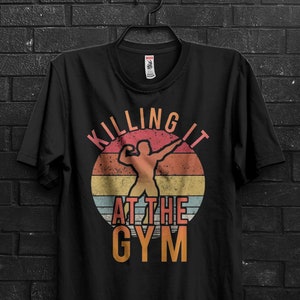 Puede incluir: Camiseta negra con un diseño retro que presenta la silueta de un levantador de pesas dentro de un círculo colorido. El texto "KILLING IT AT THE GYM" está impreso encima y debajo del gráfico en un estilo vintage.