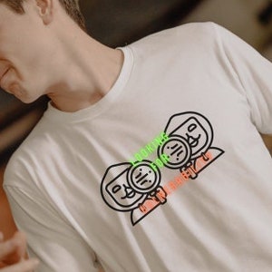 Pode incluir: Camiseta branca com um desenho animado preto e vermelho de duas figuras com detalhes verdes. A pessoa está usando shorts xadrez.