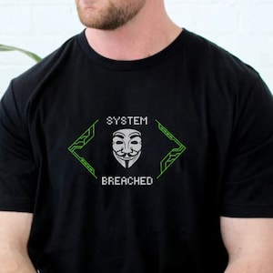 May include: Black t-shirt with a white Guy Fawkes mask design inside a green geometric frame. The words "SYSTEM BREACHED" are above and below the mask. The shirt has a crew neck and short sleeves.