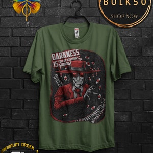 May include: Olive green t-shirt featuring a graphic of a character in a red suit and hat, surrounded by a dark, eye-filled design. The text "DARKNESS IS FULL OF MYSTERIES & SURPRISES" is displayed on the shirt.