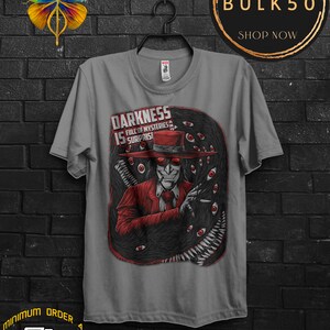 May include: Gray t-shirt featuring a graphic of a character in a red suit and hat, surrounded by black tentacles and red eyes. The text "DARKNESS IS FULL OF MYSTERIES & SURPRISES" is printed above the character. A black hanger is holding the shirt.