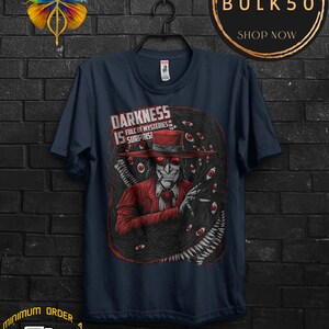 May include: Navy blue t-shirt featuring a detailed illustration of a character in a red suit and hat, surrounded by eyes and dark, swirling shapes. The text "DARKNESS IS FULL OF MYSTERIES & SURPRISES" is printed on the shirt.