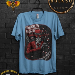 May include: Light blue t-shirt featuring a graphic of a character in a red suit and hat, surrounded by a dark, eye-filled design. Text on the shirt reads "DARKNESS IS FULL OF MYSTERIES & SURPRISES".