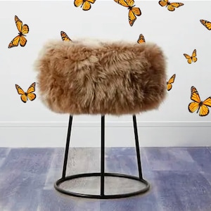 Puede incluir: Un taburete marrón y esponjoso con patas de metal negro. El taburete tiene una base redonda y tres patas. El fondo presenta una pared blanca decorada con calcomanías de mariposas naranjas y negras.