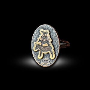 Puede incluir: Anillo ovalado con una banda y una cara texturizadas en tono plateado. La cara presenta un diseño en tono dorado y metal oscuro de un caballo que lleva tres figuras. El anillo está sobre un fondo negro.