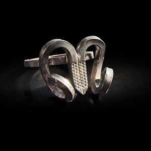 May include: A silver-toned ring with an abstract design, featuring curved shapes and a textured central element. The ring is set against a black background, highlighting its unique form and metallic sheen. The ring's design is modern and artistic.