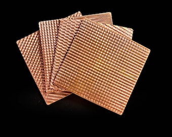 May include: Close-up of several square copper coasters with textured surfaces. The coasters have different patterns, including diagonal lines and a grid design. The copper has a warm, reddish-brown hue. The text "Elementally Copper" is at the bottom.