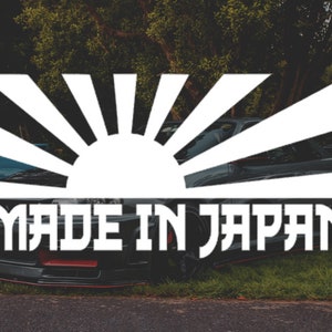 Puede incluir: Una pegatina blanca con un gráfico de sol naciente y el texto "MADE IN JAPAN" en un coche negro.