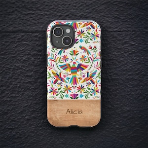 May include: A white phone case with a colorful floral and bird design. The case has a wood panel on the bottom with the name "Alicia" engraved on it.