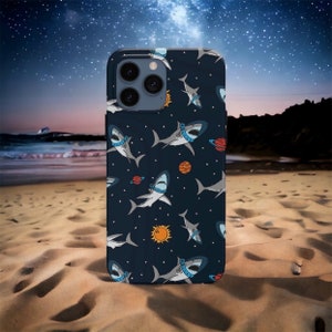 Puede incluir: Una funda de teléfono azul oscuro con un patrón de tiburones blancos con cascos espaciales y planetas. La funda tiene un recorte para la cámara con tres lentes.