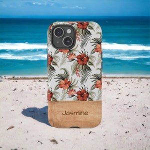 May include: A white phone case with a floral pattern of red hibiscus flowers and green leaves. The case has a wood accent at the bottom with the word "Jasmine" printed on it.