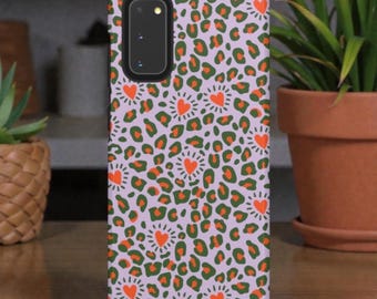 May include: A phone case with a lavender background featuring a repeating pattern of green and orange leopard spots and orange hearts with white rays. The case has a camera cutout and a black border.